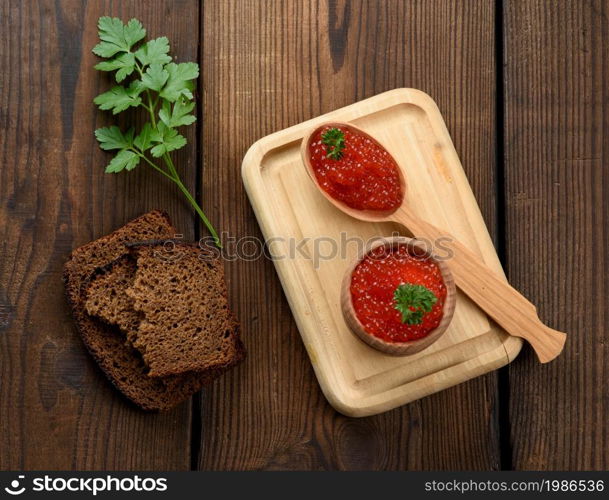 red caviar of pink salmon lies in a wooden spoon on a cutting board. Brown wooden table, top view