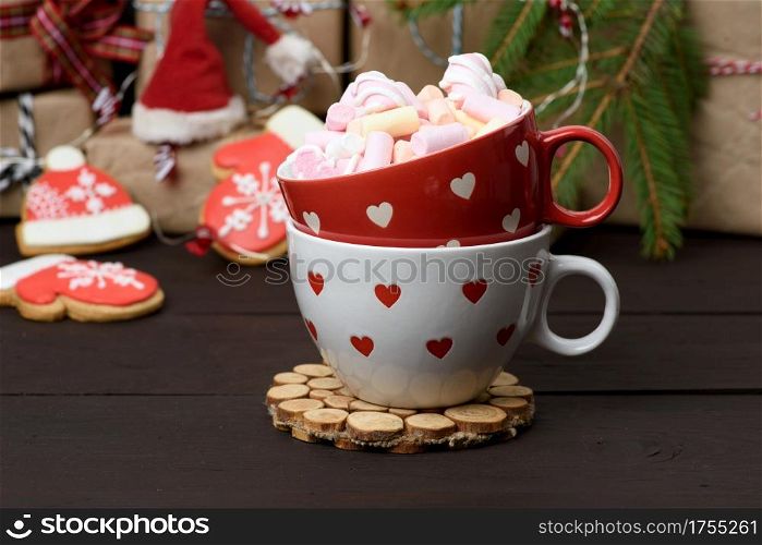 red ceramic cup with cocoa and marshmallows, behind a gift box and a Christmas garland, close up