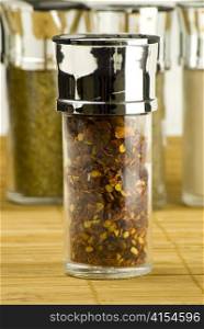 red chili flakes in a glass jar on different spices background over wooden mat