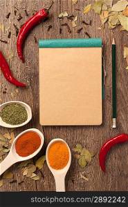Red chili peppers, spices in spoons, notebook paper and pencil on oak wood texture background