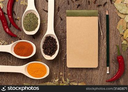 Red chili peppers, spices in spoons, notebook paper and pencil on oak wood texture background