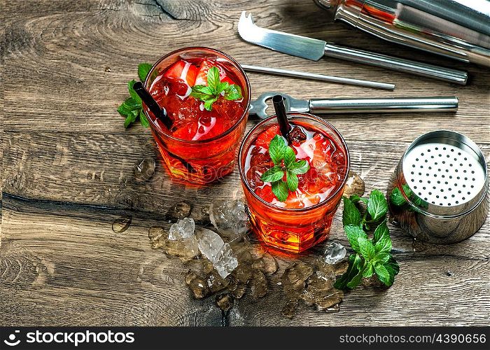 Red cocktail with strawberry, mint leaves, ice. Drink making bar accessories on wooden table