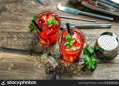 Red cocktail with strawberry, mint leaves, ice. Drink making bar accessories on wooden table