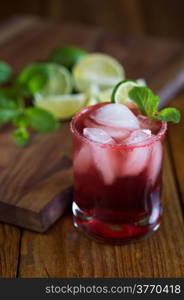 Red colored cocktail in a short glass on a rustic table garnished with limes and mint leaves.&#xA;