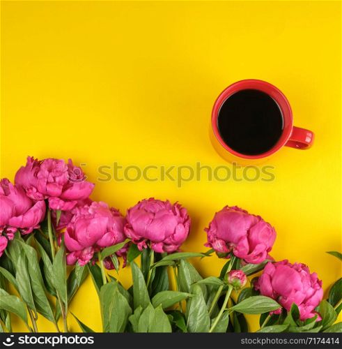 red creameric cup with black coffee and a bouquet of red peonies with green leaves on a yellow background, top view