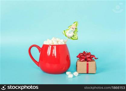 Red cup of marshmallow with green lollipop christmas tree and gift box on blue background. Merry Christmas or Happy New Year concept. Copy space Template for your design, Greeting card.. Red cup of marshmallow with green lollipop christmas tree and gift box on blue background. Merry Christmas or Happy New Year concept. Copy space Template for your design, Greeting card