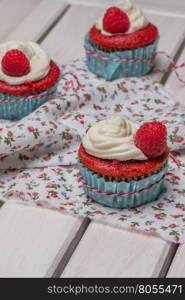 Red cupcakes with cream cheese frosting and raspberries on white wooden table.