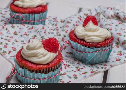Red cupcakes with cream cheese frosting and raspberries on white wooden table.