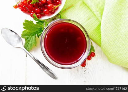 Red currant jam in a glass jar, bunches of berries with leaves, a napkin and a spoon on wooden board background from above