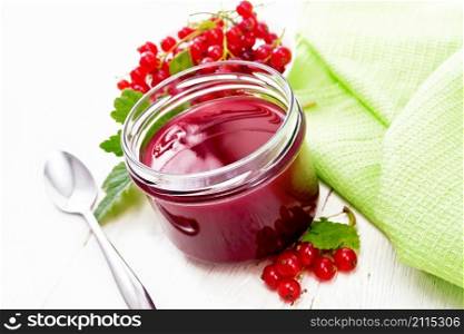 Red currant jam in a glass jar, bunches of berries with leaves, a napkin and a spoon on wooden board background