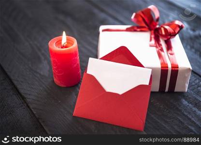Red empty envelope leaned against a stylishly wrapped gift, tied with red ribbon and bow, near a lit candle on an old wooden table.