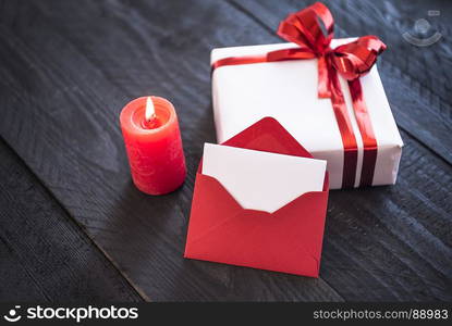 Red envelope with blank paper near a lit candle and a gift box wrapped in white paper and tied with red ribbon and bow.