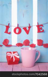 Red gift box near a pink cup with the word love spelled from red paper letters, tied to a string and soap rose petals on a blue wooden fence.