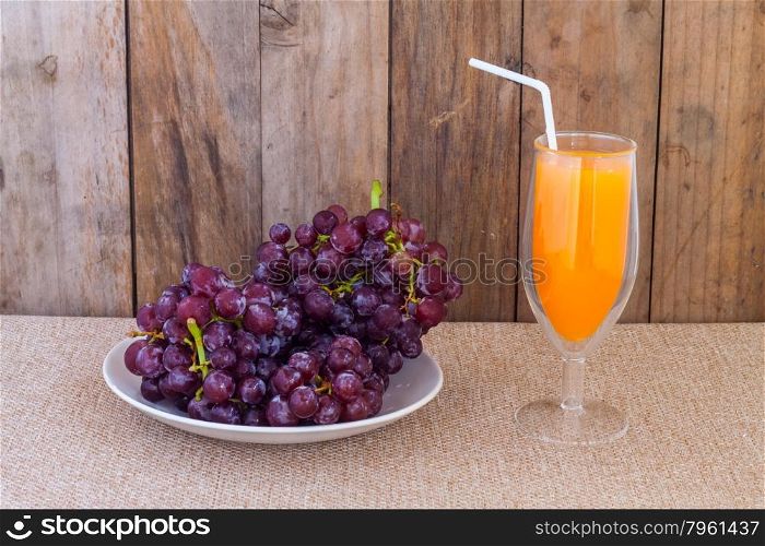 Red grapes in dish and orange juice in glass with white straw against wooden background