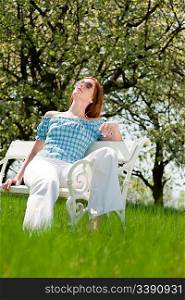 Red hair woman enjoying spring sun on white bench in a meadow; shallow DOF