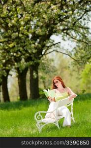 Red hair woman reading book on white bench in green meadow, shallow DOF