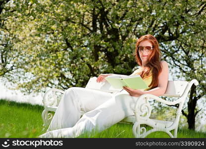 Red hair woman reading book on white bench in green meadow, shallow DOF