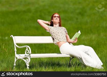 Red hair woman sitting with book on white bench in a meadow; shallow DOF
