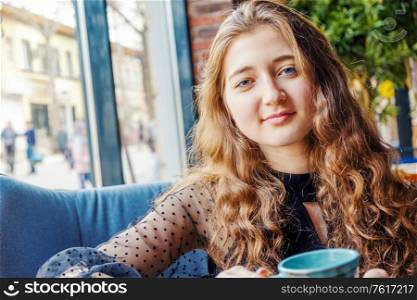 Red-haired girl with blue eyes sits in a cafe and looks into the camera over the table with cups. Red-haired girl with blue eyes sits in a cafe and looks into the camera