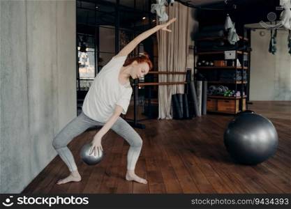 Red-haired woman in gym, doing plie with fitball in hand, embraces sporty ambiance. Fitness activities showcased.