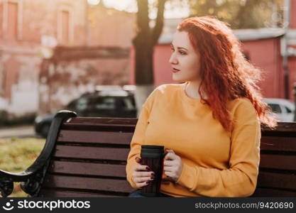 Red-haired woman in yellow sweatshirt sits on park bench and drinks coffee. She is holding reusable mug in her hands. Portrait on sunny autumn day. Lifestyle.. Red-haired woman in yellow sweatshirt sits on park bench and drinks coffee. She is holding reusable mug in her hands. Portrait on sunny autumn day.