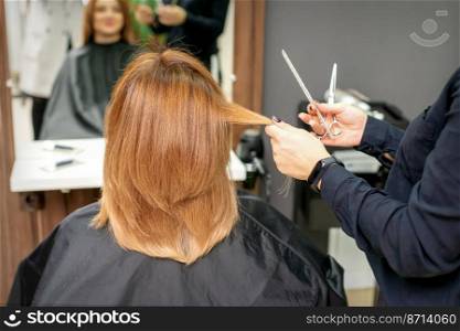 Red-haired woman sitting a front of the mirror and receiving haircut her red long hair by a female hairdresser in a hair salon, back view. Red-haired woman sitting a front of the mirror and receiving haircut her red long hair by a female hairdresser in a hair salon, back view.