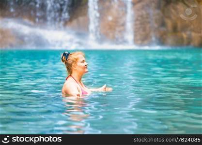 Red haired young woman in pink bikini swimsuit relaxes in emerald tropical lake with waterfall. Erawan National park, Kanchanaburi, Thailand
