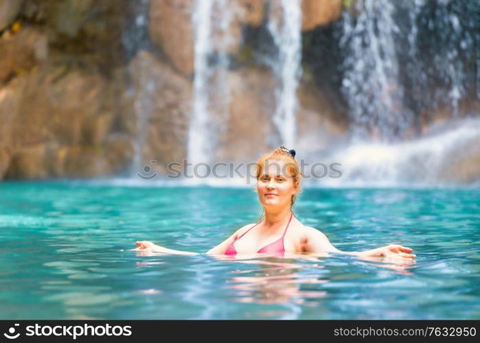 Red haired young woman in pink bikini swimsuit relaxes in emerald tropical lake with waterfall. Erawan National park, Kanchanaburi, Thailand