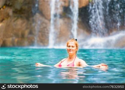 Red haired young woman in pink bikini swimsuit relaxes in emerald tropical lake with waterfall. Erawan National park, Kanchanaburi, Thailand