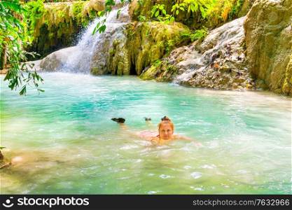 Red haired young woman in pink bikini swimsuit swims in emerald blue water of tropical lake with waterfall. Erawan National park, Kanchanaburi, Thailand