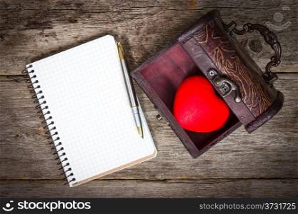 Red heart in the box and notebook with pen on wooden floor