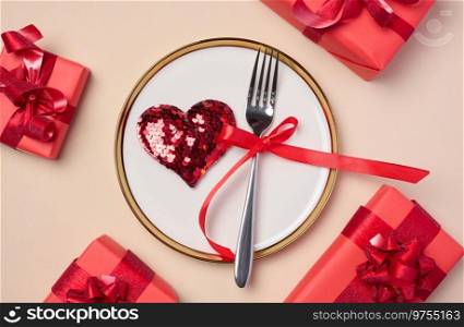 Red heart on a round white plate and fork on a beige background, top view. Gifts all around