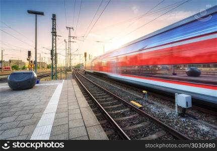 Red high speed train in motion on the railway station at colorful sunset. Germany. Blurred modern intercity train on the railway platform. Passenger train on railroad. Railway travel in Europe. Dusk. Red high speed train in motion on the railway station