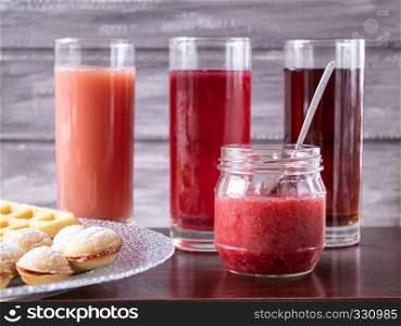 Red juice in a glass near a bowl of biscuits and a jar of jam.. Red juice in a glass next to a bowl of cookies and a small jar of jam.