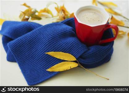 red mug with coffee on the table in a blue knitted scarf and yellow autumn leaves. hello autumn concept. seasonal diseases