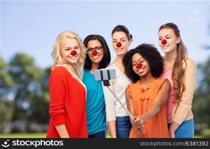 red nose day and people concept - international group of happy smiling different women with clown noses taking picture with smartphone over natural background. group of women taking selfie at red nose day