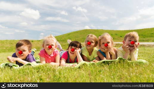 red nose day, charity and childhood concept - group of happy kids with clown noses lying on blanket or cover on meadow. happy kids lying on meadow at red nose day