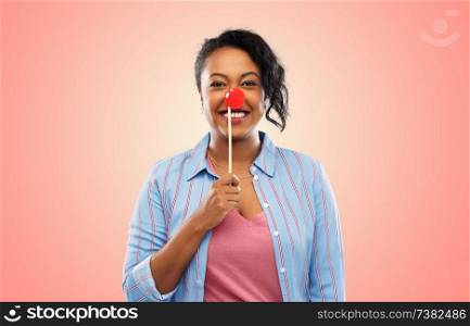 red nose day, party props and photo booth concept concept - happy african american young woman with clown nose over living coral background. happy african american woman with red clown nose