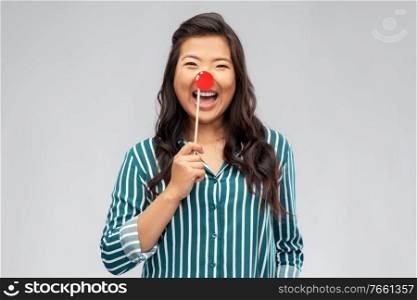 red nose day, party props and photo booth concept concept - happy asian young woman with clown nose over grey background. happy asian woman with red clown nose
