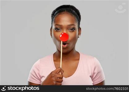 red nose day, party props and photo booth concept concept - surprised african american young woman with clown nose over grey background. surprised african woman with red clown nose