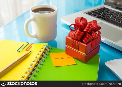 Red present box and paper note with Merry christmas message: Ho Ho Ho! place on office table with laptop, colorful diary and a cup of coffee, concept to christmas gift and holiday season