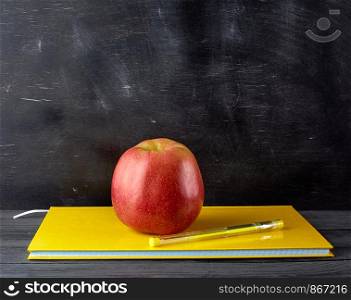 red ripe apple lies on a yellow closed notebook, background from black chalk board