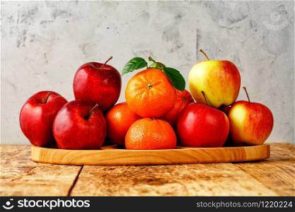 Red ripe apples and mandarins with green leaves lie on a wooden tray on an old wooden table on a gray concrete background, image with copy space.. Red ripe apples and tangerines with green leaves lie on a wooden tray on an old wooden table with gray concrete background.