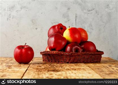 Red ripe apples lie in a wicker basket on an old wooden table on a gray concrete background, image with copy space.. Red ripe apples lie in a wicker basket on an old wooden table with gray concrete background.