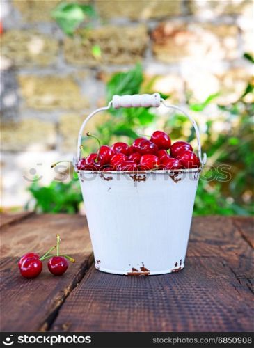 Red ripe cherry in a white iron bucket on a brown wooden table. Red ripe cherry in a white iron bucket