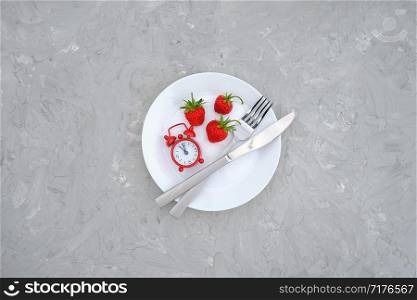 Red ripe strawberries berry on white plate, cutlery and red alarm clock on gray stone background table. Top view, flat lay, copy space Template. Concept diet and detox time or summer menu time.. Red ripe strawberries berry on white plate, cutlery and red alarm clock on gray stone background table. Top view, flat lay, copy space Template. Concept diet and detox time or summer menu time