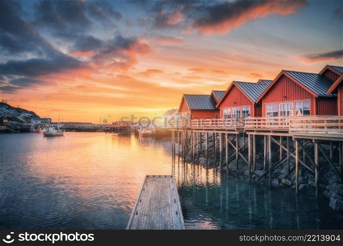 Red rorbu on wooden piles on sea coast, small jetty, colorful orange sky at sunrise in winter. Lofoten islands, Norway. Traditional norwegian rorbuer, reflection in water, boats in fishing village