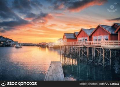 Red rorbu on wooden piles on sea coast, small jetty, colorful orange sky at sunrise in winter. Lofoten islands, Norway. Traditional norwegian rorbuer, reflection in water, boats in fishing village