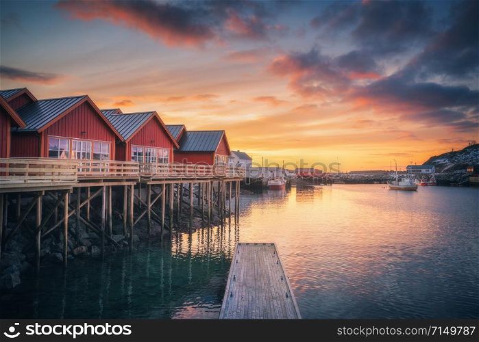 Red rorbu on wooden piles on sea coast, small jetty, colorful sky at amazing sunrise in winter. Lofoten islands, Norway. Traditional norwegian rorbuer, reflection in water, boats in fishing village