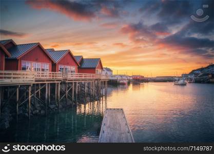 Red rorbu on wooden piles on sea coast, small jetty, colorful sky at amazing sunrise in winter. Lofoten islands, Norway. Traditional norwegian rorbuer, reflection in water, boats in fishing village